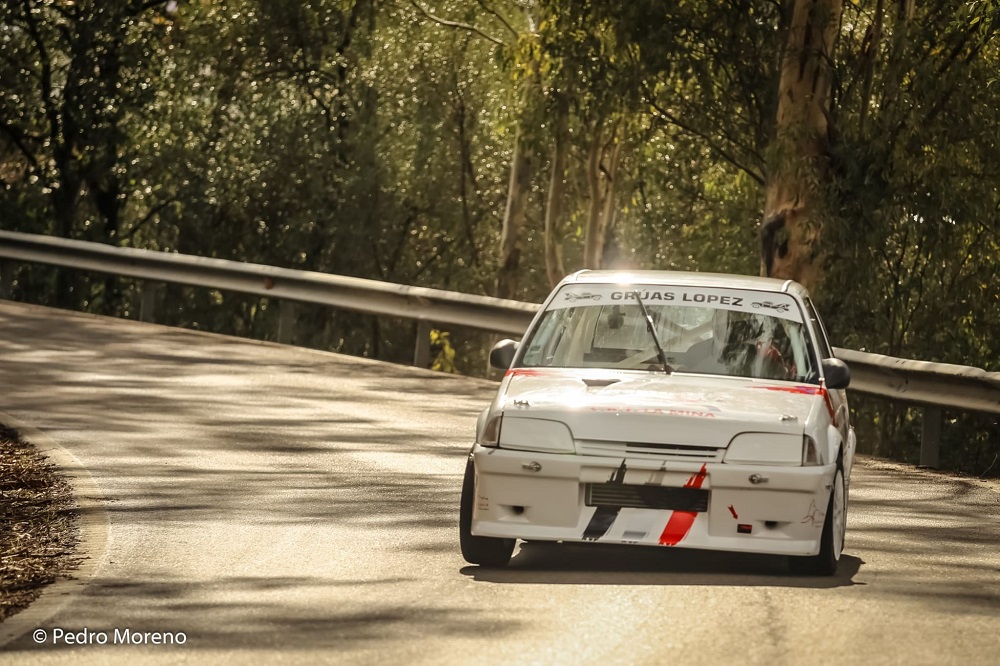 A white Citroen AX race car, driven by Antonio Zambrana, speeding uphill on a mountain road during a rally event, with a green forest in the background.